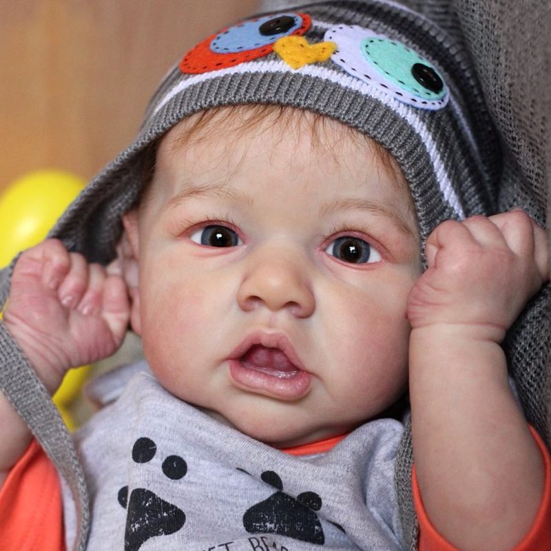 A baby with big eyes wears a hat with an owl design and a shirt with bear prints, looking directly at the camera.