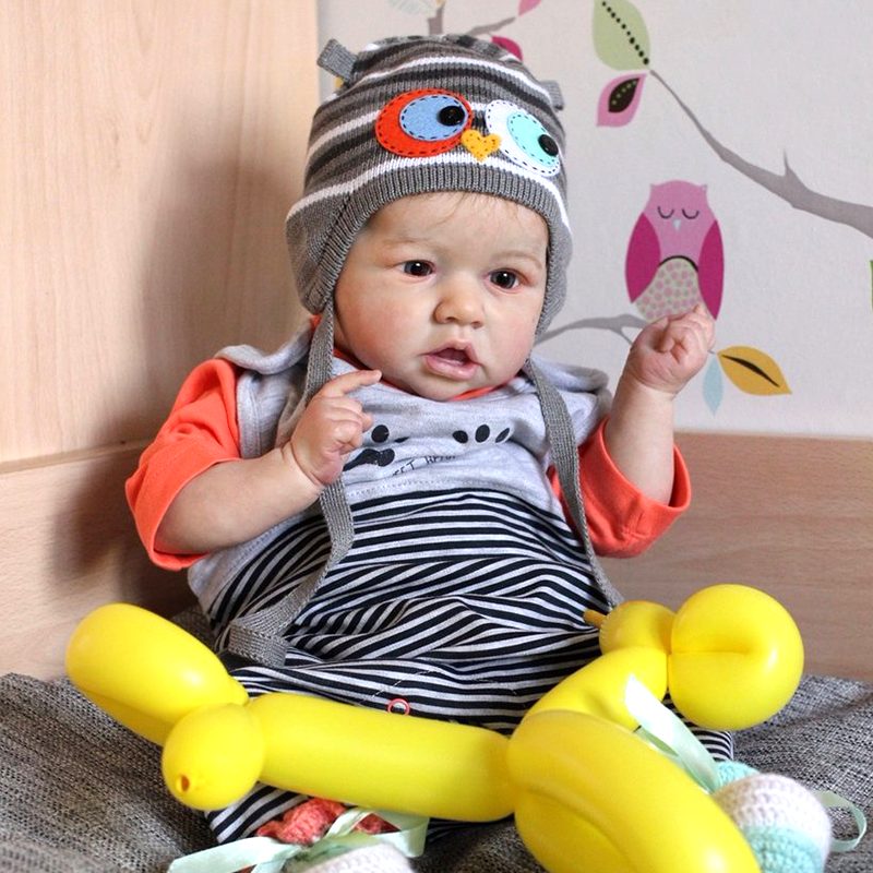 A baby wearing an owl hat, orange sleeves, and striped pants, sitting with a yellow balloon animal.