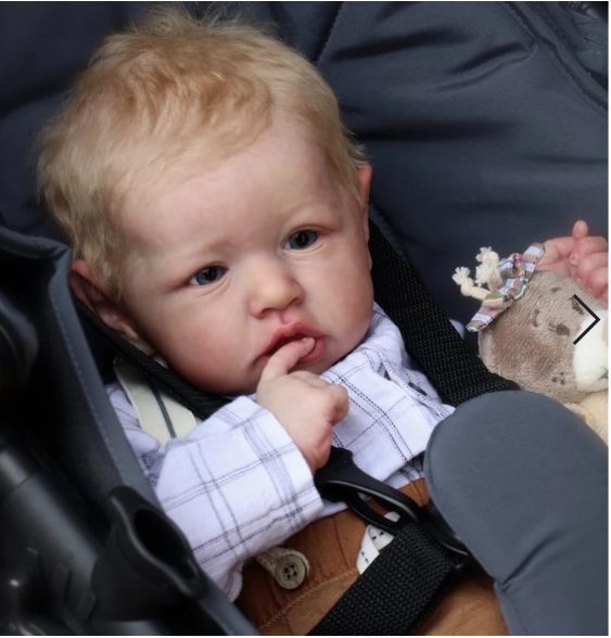 A baby with blonde hair and blue eyes, sitting in a stroller with a finger in their mouth and a stuffed toy next to them, looks just like one of those realistic dolls from a top doll supplier.