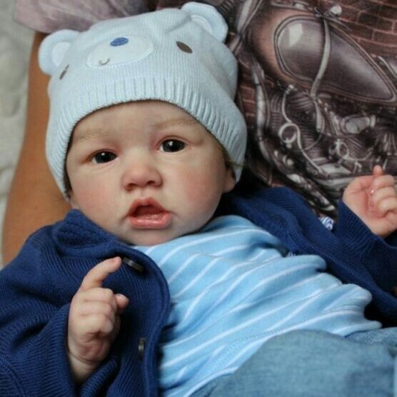 A reborn baby girl in a blue bear hat and blue striped outfit lies on someone's lap, looking towards the camera.