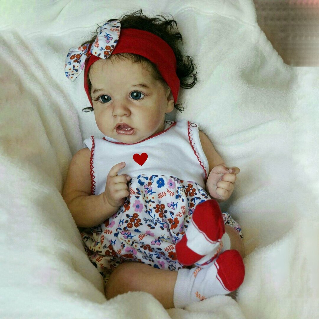 A baby with curly hair wearing a red headband, white and floral dress, and red shoes sits on a white blanket, looking as charming as the realistic dolls crafted by a skilled doll supplier.