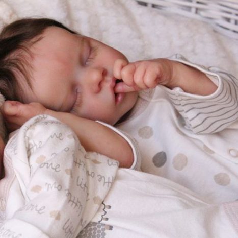A reborn baby girl peacefully sleeping, wearing a white onesie with polka dots, and holding her hand near her face.