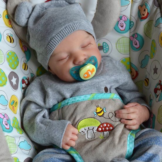A sleeping reborn baby girl in a gray hat and matching outfit with a pacifier, resting in a colorful, patterned baby seat.