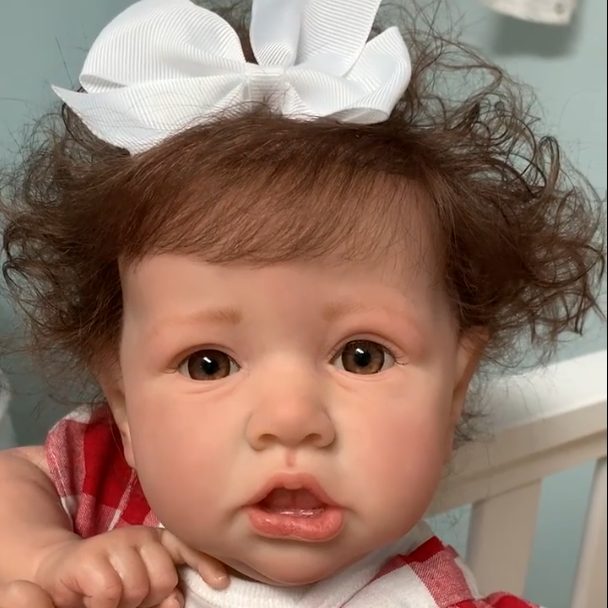 A lifelike baby doll with curly hair, a white bow, and a red-checkered outfit. This realistic reborn doll captures the innocence and charm of a real infant.