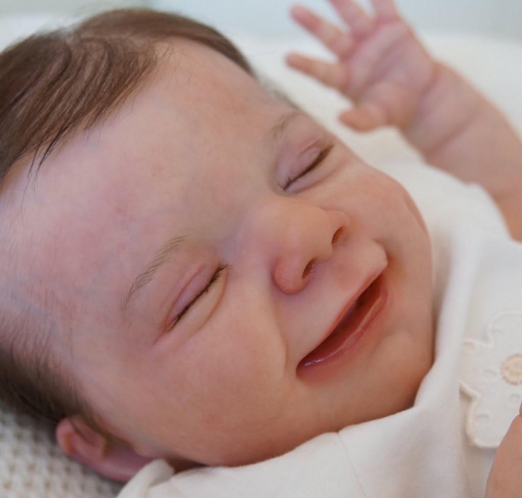 A close-up of a lifelike reborn doll with eyes closed, smiling, and raising one hand.