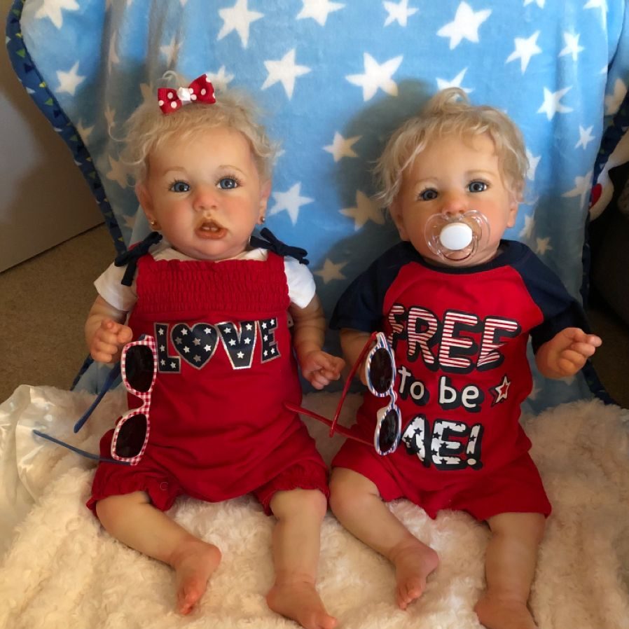 Two reborn dolls in patriotic clothing sit on a blue star-patterned blanket, each holding sunglasses, one with a pacifier.