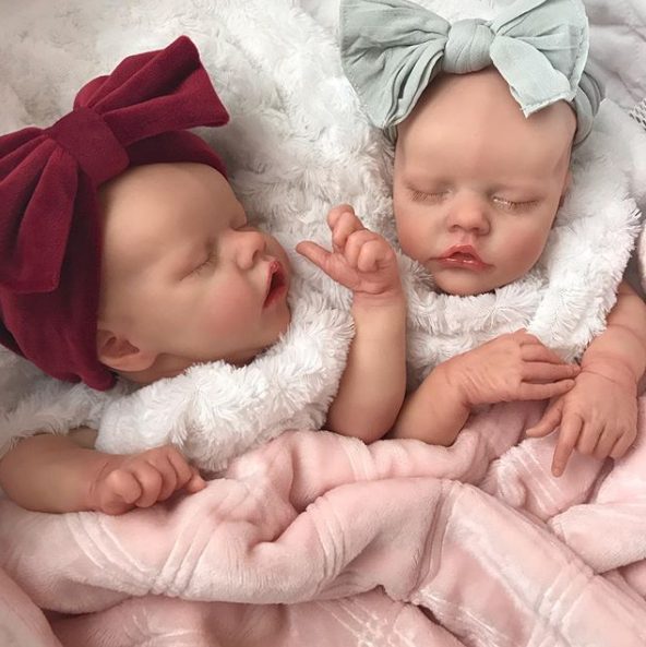 Two realistic dolls with headbands cuddle under a fluffy, pink blanket.